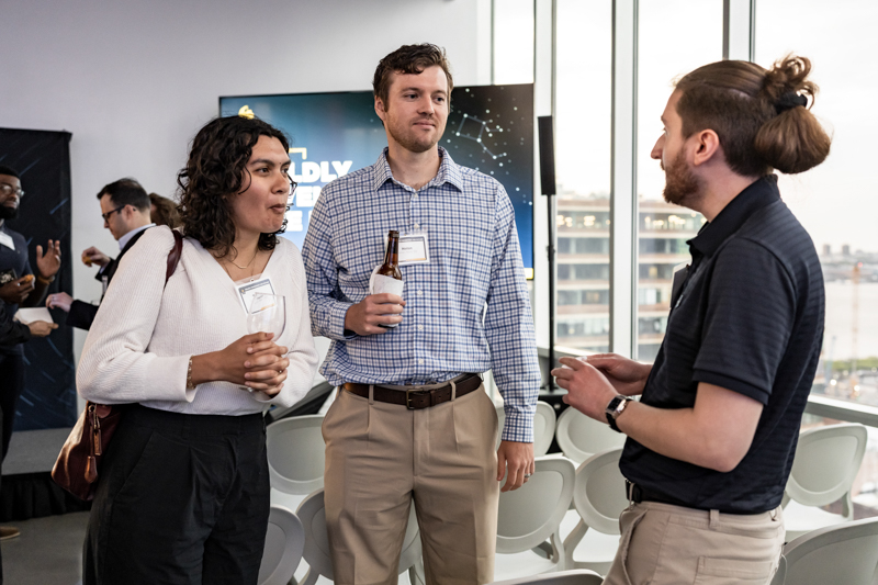 Three professionals chat at a networking event; one holds a beer bottle while a presentation screen and large windows loom in the background.