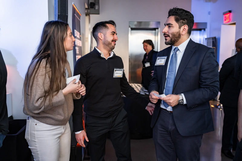 Three professionals at a networking event chatting by a wall with name badges visible, woman on the left listening as two men speak.