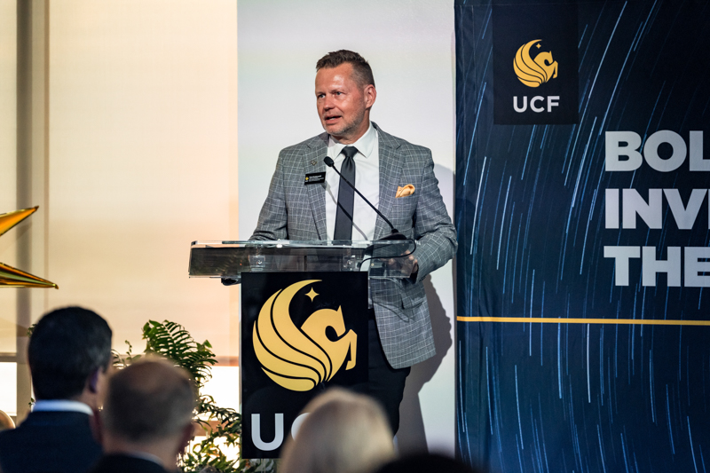 Man in a grey plaid blazer speaks at a lectern with a microphone during a UCF event.