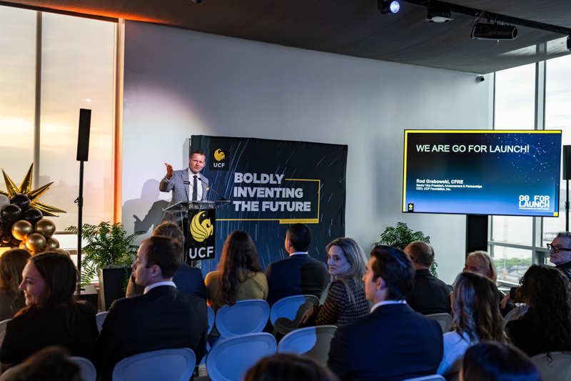Speaker at podium presenting to an audience, with a banner reading Boldly Inventing the Future and a screen saying We are go for launch! (UCf branding)