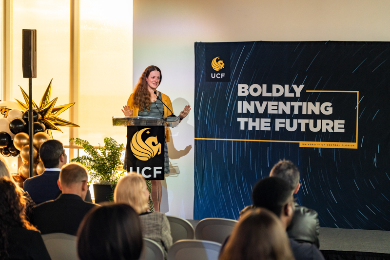 Woman speaks at a podium during a UCF event, with a large banner reading 'Boldly Inventing the Future' in the background and seated audience in the foreground.