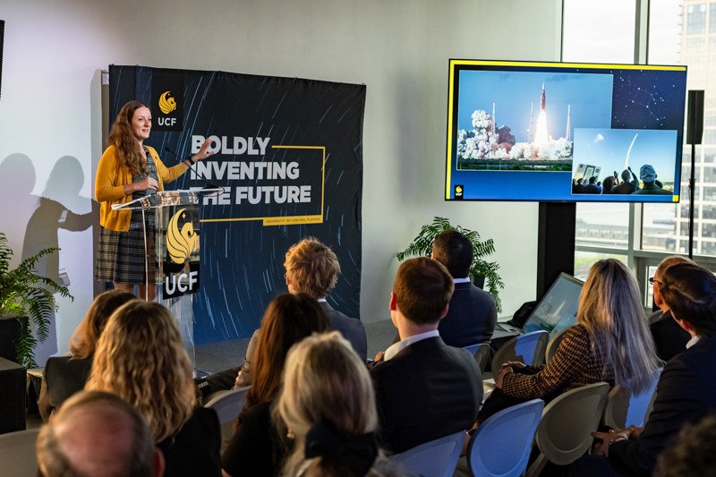 Woman in a mustard cardigan speaks at a podium during a conference titled 'Boldly Inventing the Future' with a rocket image on the screen behind her.