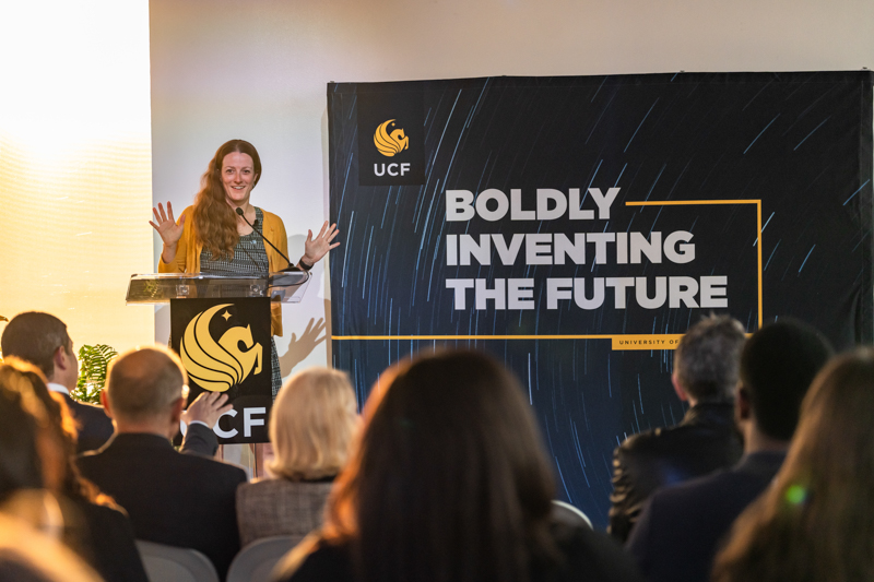 Woman speaking at a podium in front of a banner that reads 'Boldly Inventing the Future' with the UCF logo visible.