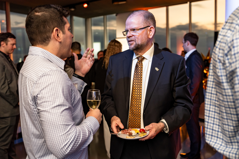 Two men at a business reception chatting; one holds a glass of white wine, the other a plate of appetizers.