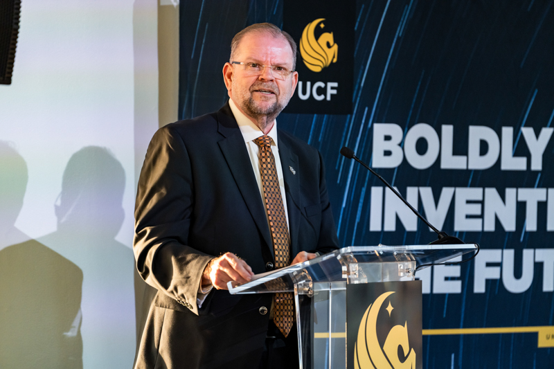 Man in a suit speaks at a clear podium during a conference; backdrop features the UCF logo and large 'BOLDLY INVENT...' text.