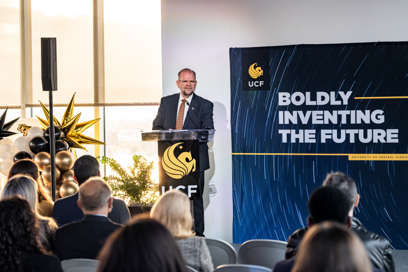 Man in a suit giving a keynote at a UCF event, standing at a podium with the UCF logo and a banner reading 'Boldly Inventing the Future'.