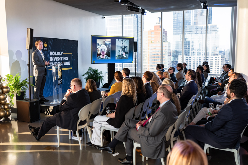 Business speaker on a stage presenting to a seated audience in a modern office, with a video-call screen in the background.