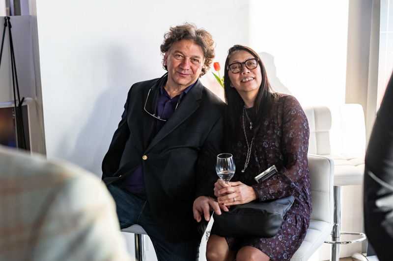 Two smiling adults sit close together on white chairs, posing for the camera at a social event with a wine glass nearby.
