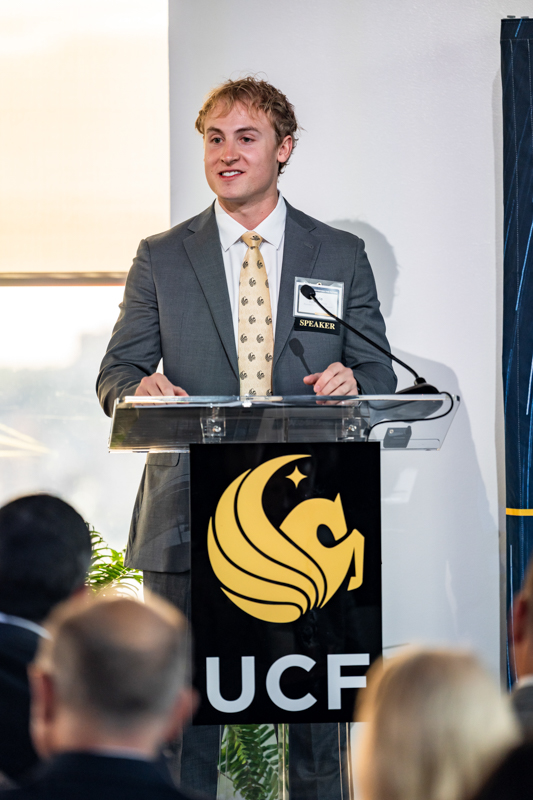 Speaker at a clear lectern delivering a talk, wearing a gray suit and patterned tie, with a 'SPEAKER' badge and a large UCF emblem on the banner behind.