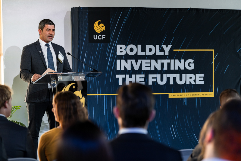 Man in a dark suit speaks at a clear podium beside a banner that reads 'Boldly Inventing the Future' with the UCF logo in the corner, during a formal event.Audience seen in foreground.