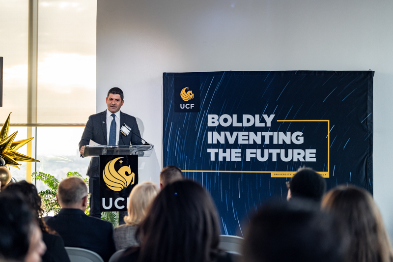 Male speaker at a lectern with the UCF logo, addressing an audience beside a banner that says Boldly Inventing the Future.