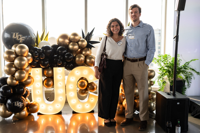 Two adults posing at a UCF event behind a gold and black balloon arch and lit 'UCF' letters on display.