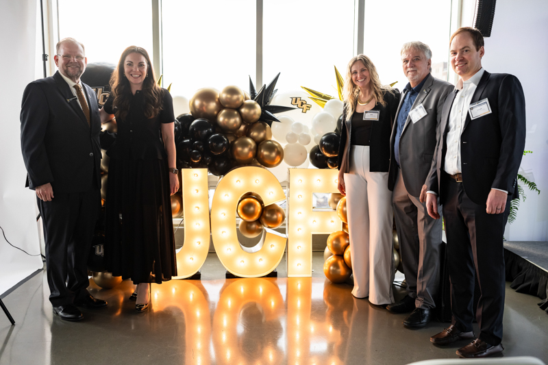 Six professionally dressed people posing behind illuminated JCH letters, with gold, black, and white balloons in a photo studio.