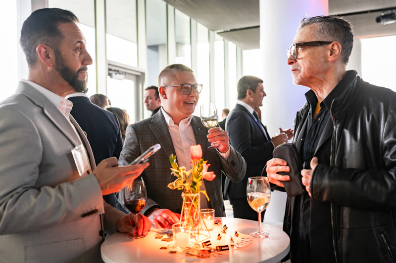 Three professionals mingle at a round cocktail table, toasting with wine glasses at a networking event.