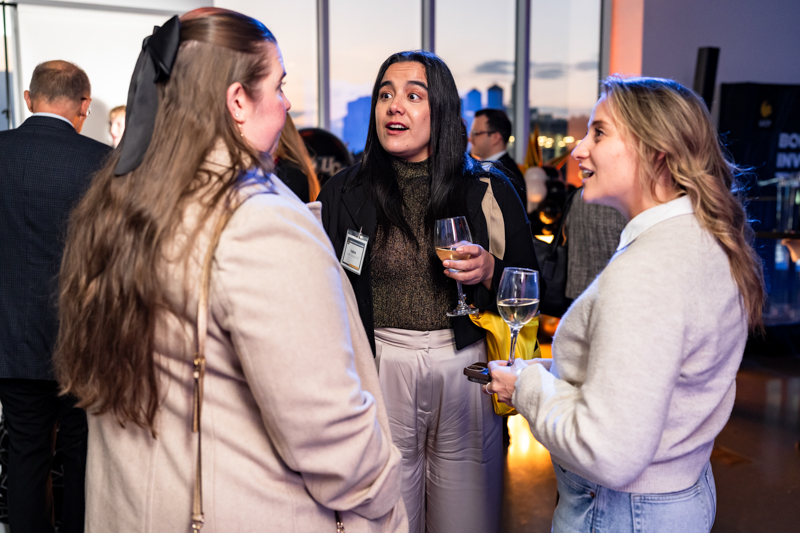 Three women at a networking event chat, each holding a wine glass, with a visible name badge on the center person.