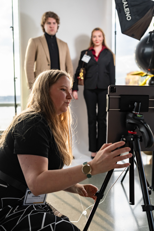 Photographer’s assistant adjusts a tablet on a tripod in a studio as two models pose in the background near lighting equipment.