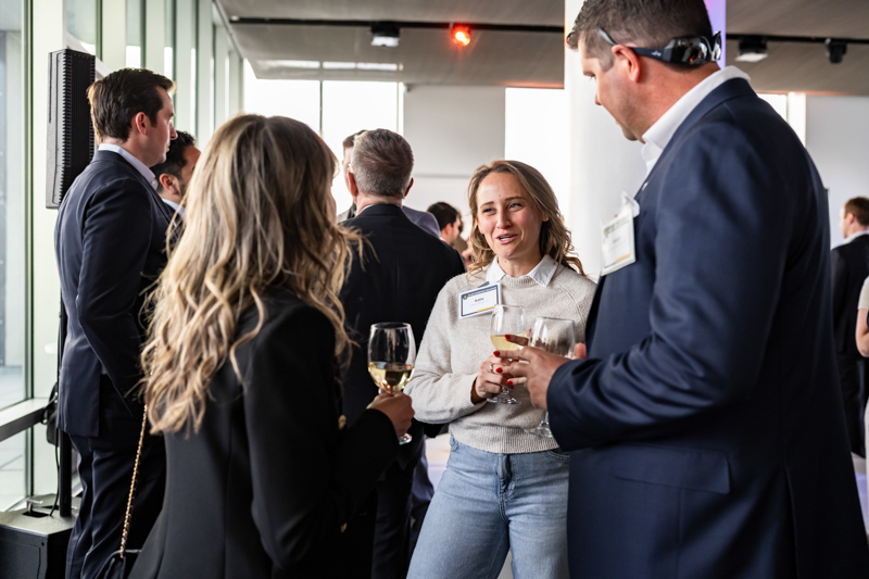 Professionals networking at a reception, holding wine glasses and wearing name badges in a modern venue near large windows.