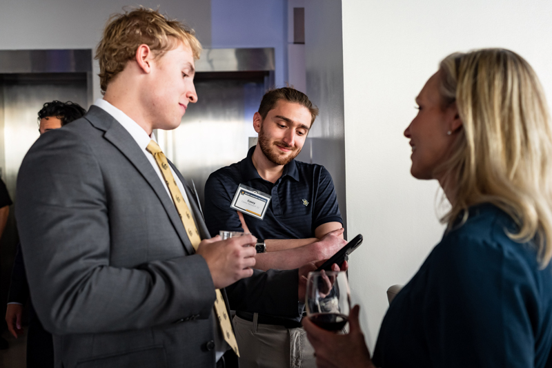 Three professionals chat at a networking event, one man in a suit holding a glass and listening to a woman.