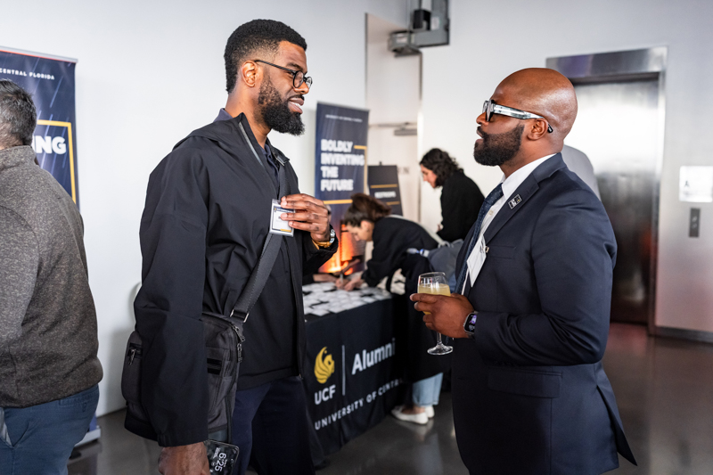 Two men chat at a university alumni event; one wears a badge and carries a bag, the other in a suit holds a wine glass near an alumni table.