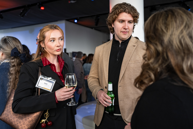Two adults at a reception talking to someone offscreen: the woman holds a wine glass and wears a name badge, the man beside her holds a beer bottle and wears a beige jacket.
