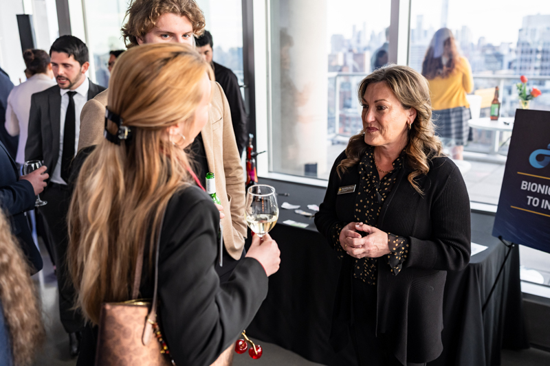 Business networking event: a woman in a black blazer talks with a guest who holds a wine glass by a table near a window.