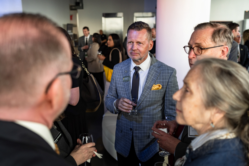 Group of business professionals mingling at a corporate reception; man in a blue checkered suit with a yellow pocket square holds a glass at the center of the frame.