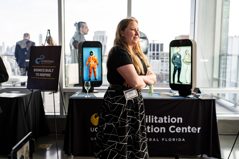 Woman with a lanyard stands at an expo booth featuring large screens displaying 3D-printed devices and an assistive-tech display (Limbitless).