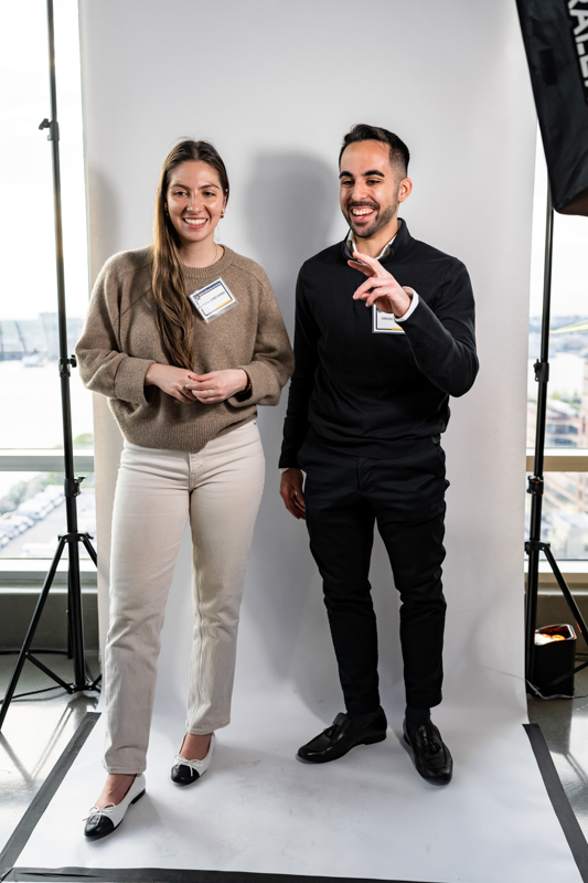 Two people stand in a photography studio with a white backdrop and visible lights; a smiling woman in a beige sweater and light pants, and a smiling man in a black outfit wearing name tags.