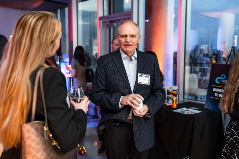 Older man in a dark blazer and light shirt talks to a woman with a wine glass at a networking event, name badge on his chest.