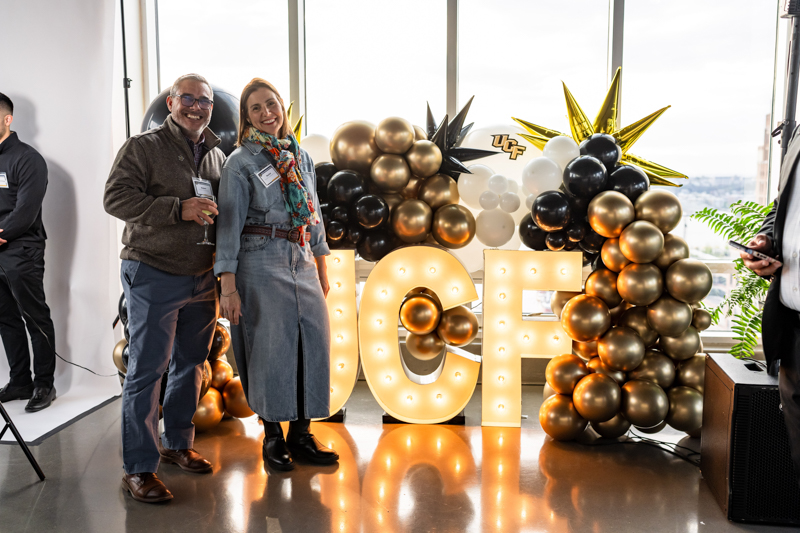 Smiling couple posing beside large illuminated letters spelling 'LOVE' and a balloon arch in a bright studio. - - - Two adults stand with drinks in front of a glowing sign at a festive event.
