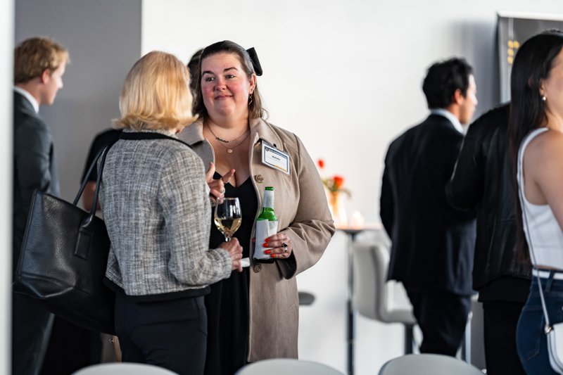 Two women in business attire chat at a networking event; one holds a wine glass and a bottle of water.