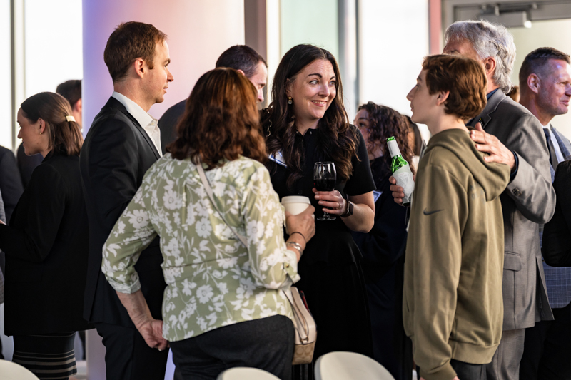People in business attire mingle at a reception, chatting and holding drinks in a bright, modern venue.