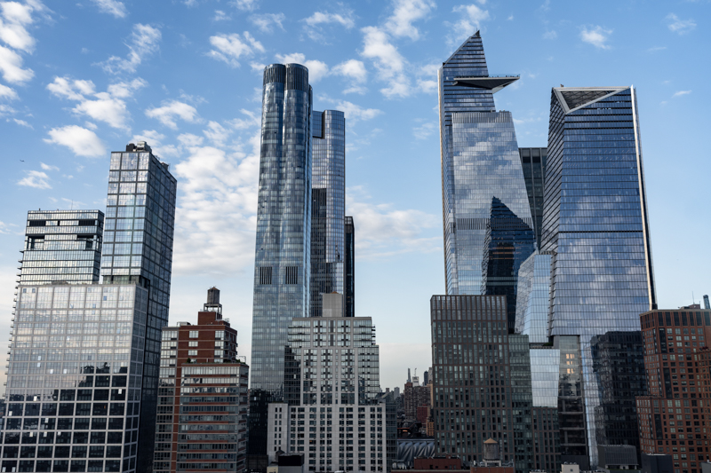 Modern city skyline with tall glass skyscrapers under a blue sky, reflecting clouds on the buildings' surfaces.