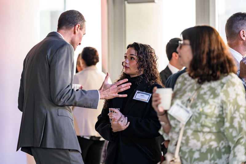 Two professionals converse in a hallway at a networking event, adults holding cups and name tags visible.