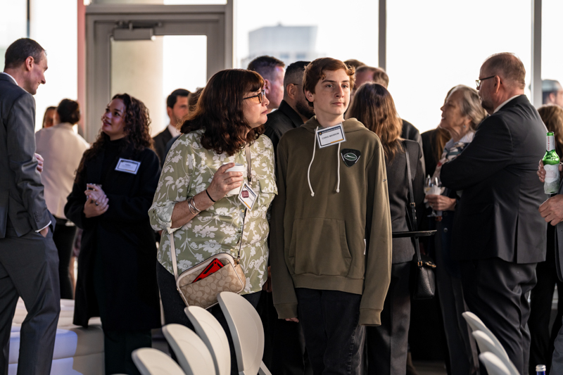 Woman in floral blouse with name badge leans toward a teenage boy in a green hoodie at a crowded reception.