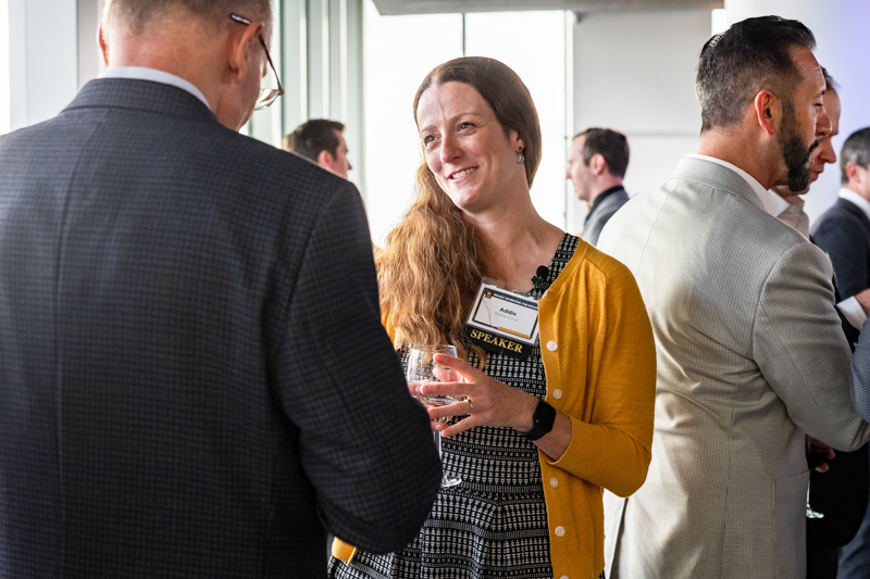Woman in a yellow cardigan chats with a man at a networking event, holding a wine glass; name tag reads SPEAKER in a lively reception setting.