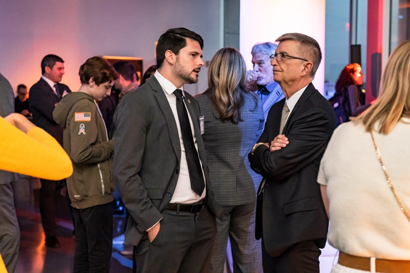 Two men in suits converse at a networking event in a modern, well-lit venue, with several people mingling in the background.