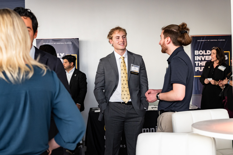 Young man in a gray suit with a name tag chats with another attendee at a networking event, backdrop banners in a university setting visible behind him
