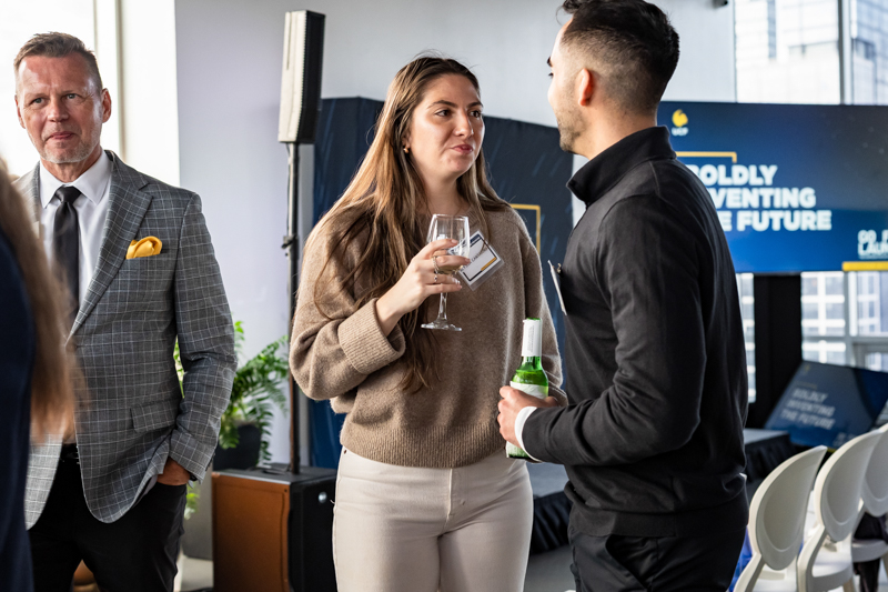 Two professionals chat at a networking event; woman holds a wine glass and badge, man in black shirt holds a beer bottle, conference space in background.