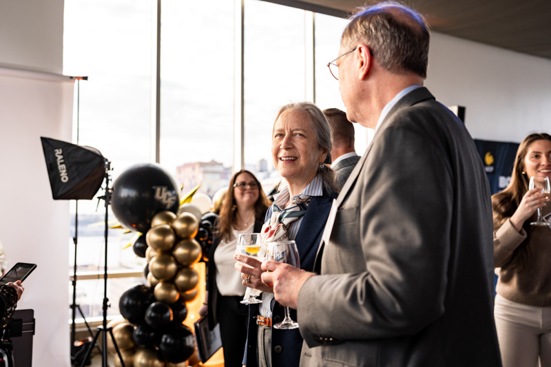 Group of professionally dressed adults at a reception, chatting and holding wine glasses near black and gold balloons.