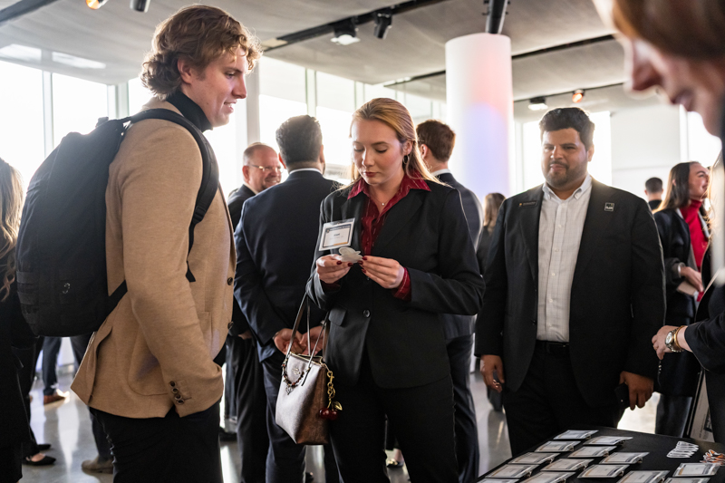 Professional networking event: a woman in a black blazer checks her name badge while a man with a backpack watches nearby in a busy lobby table setting.