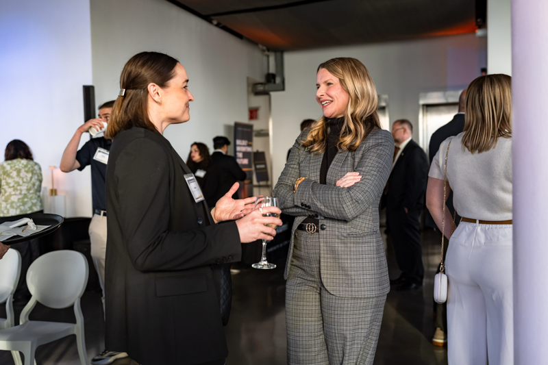 Two professionally dressed women converse at a networking event, holding wine glasses at a reception.