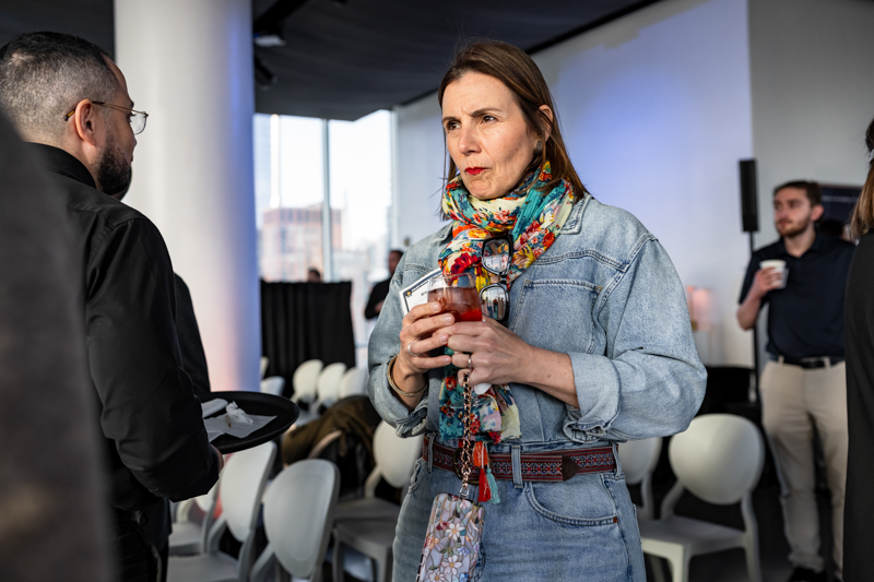 Woman in a denim jacket and colorful scarf holds a red cocktail while conversing with a man at a social event.