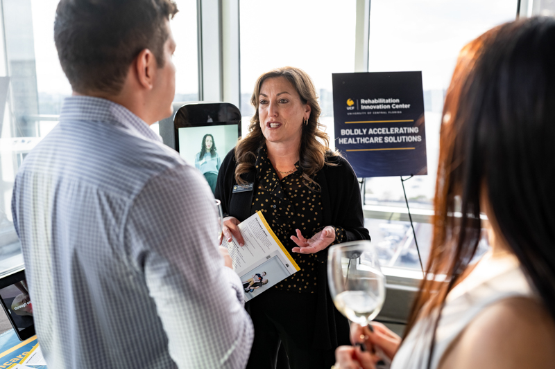 Professional woman explains a brochure to a man and a woman at a networking event, with a healthcare solutions poster in the background.