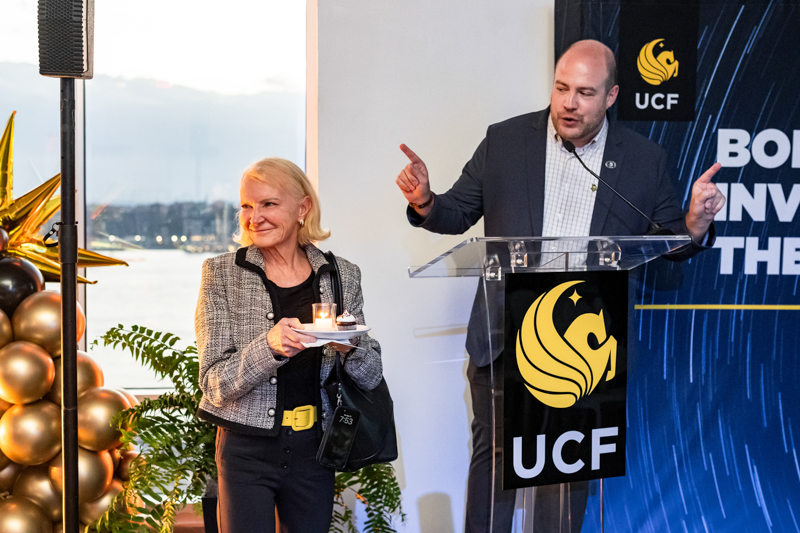 A man in a suit speaks at a clear podium with a UCF banner beside him while a smiling woman holds a lit candle and a small plate, standing near balloons by a waterfront backdrop.