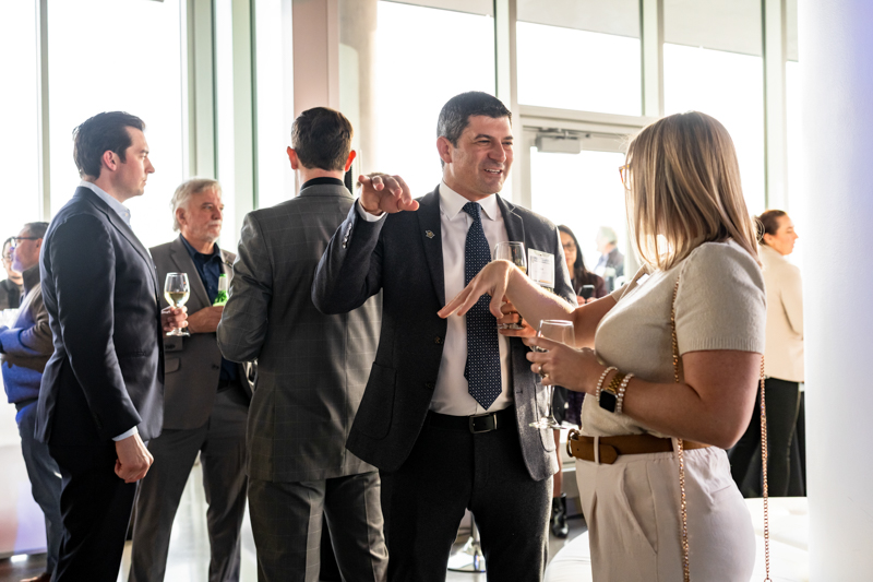 Group of professionals at a networking event, with a man in a dark suit gesturing and speaking to a woman holding a drink.