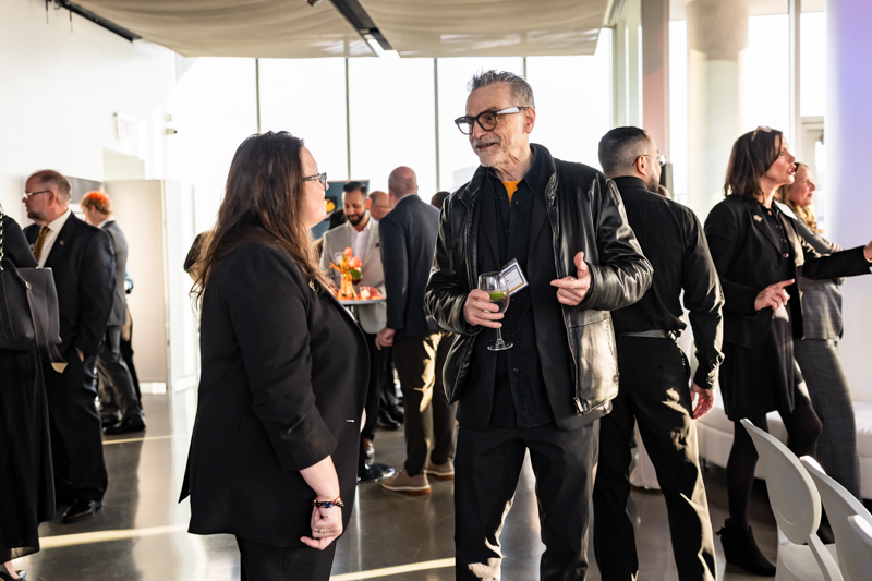 Older man in a black leather jacket chats with a woman in black at a social event, holding a wine glass and a name badge, bright room background