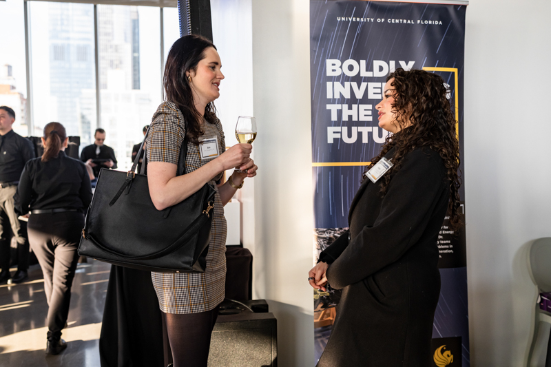 Two women in a hallway at a professional event chat, one holding a wine glass and a large black handbag, near a banner that reads 'Boldly Invest the Future'.