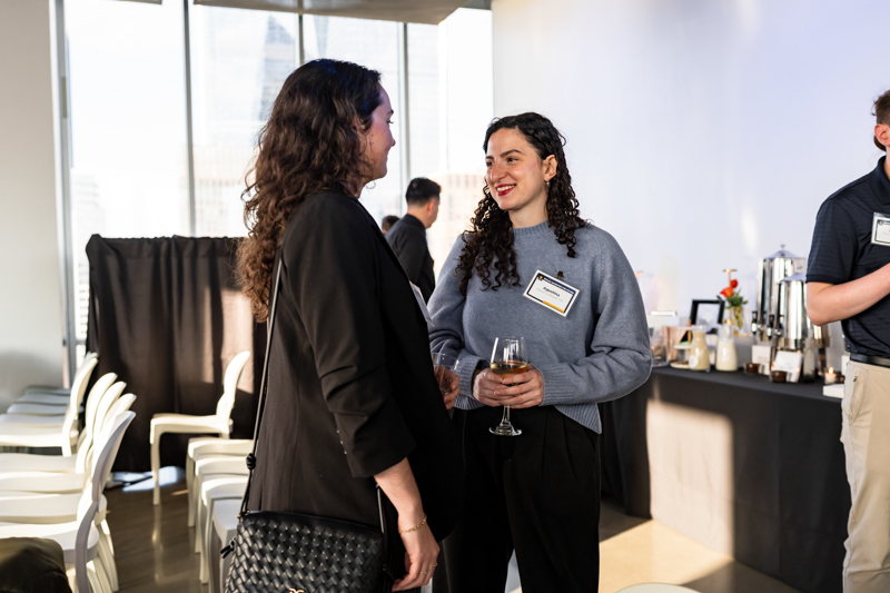 Two women in a networking event chat, one holding a glass of white wine, both wearing name badges with a refreshment table in the background.