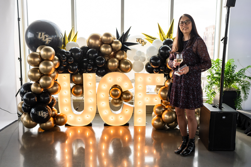 Woman in a dark floral dress with glasses standing beside a large illuminated 'UCF' sign and gold/black balloons at a festive event, holding a wine glass.